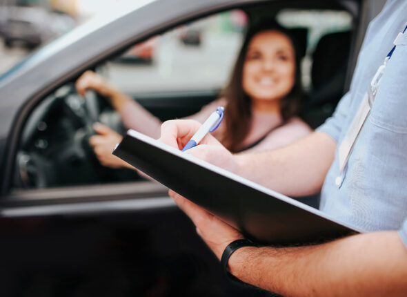 Male auto instructor takes exam in young woman. Blurred background of model sitting in car and holding hands on steering wheel. Guy hold folder with paper in hands.